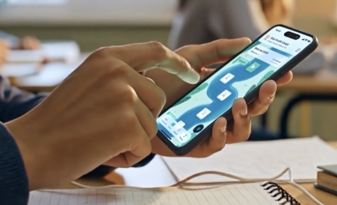 A student's hands holding a phone that shows their venture's progress map, with a notebook on the desk.